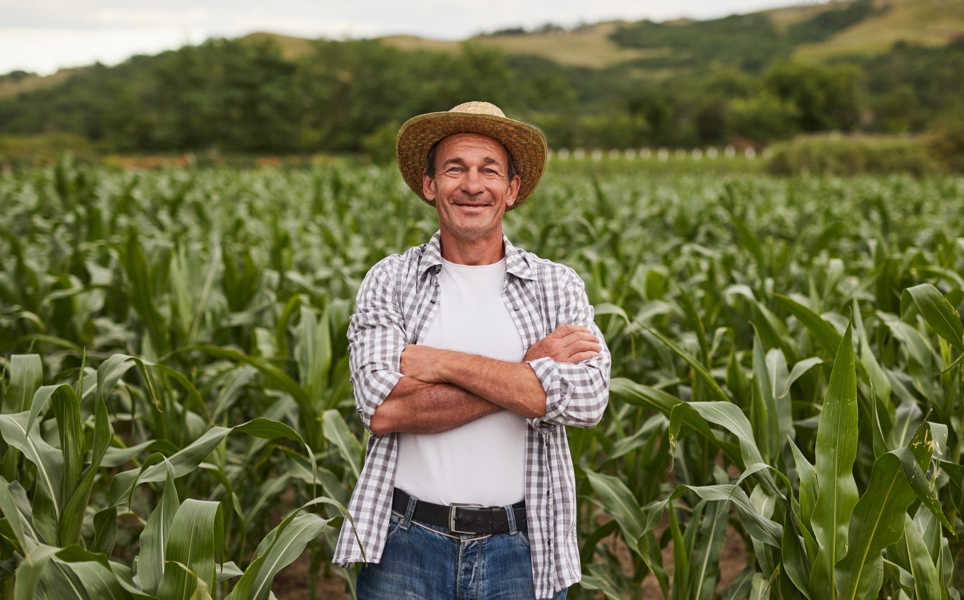 Farmer standing in a green crop field, smiling with arms crossed under a clear sky.